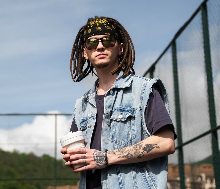 Young man with tattoos and sunglasses wearing a denim vest holding a cup outdoors near a fenced sports court, school incidents theme.