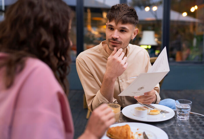 Young man at a café looking confused while discussing relationship dealbreakers with a woman during a casual meal outdoors.