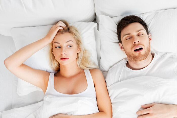 Woman looking annoyed next to a sleeping man in bed, illustrating absurd dealbreakers in relationships men are clueless about.