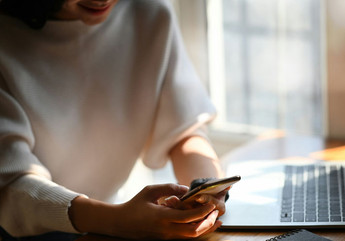 Persona con camiseta blanca usando teléfono móvil junto a laptop, ilustrando cómo los teléfonos ayudan a descubrir verdades ocultas.