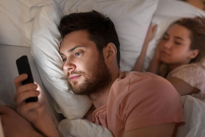 Man lying in bed using phone while partner sleeps, illustrating discovering partners' secret stories online.