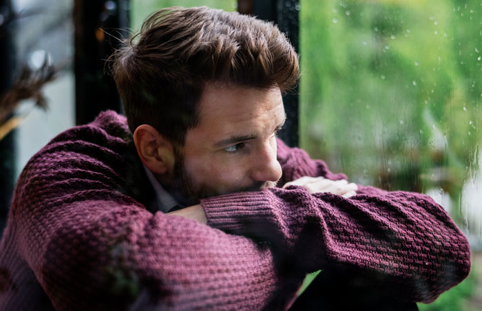 Young man in a maroon sweater looking out a window, reflecting on family expectations and caring for autistic brother.