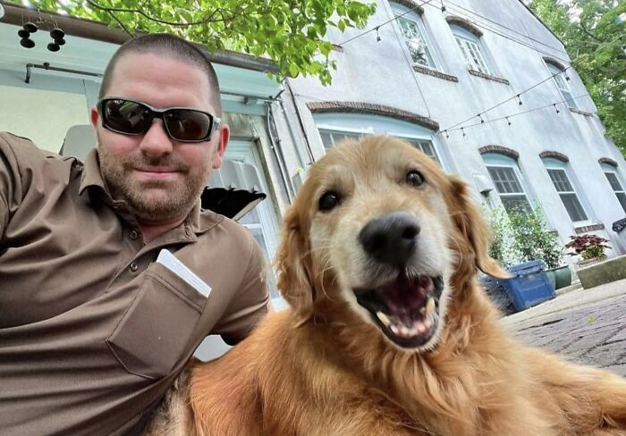 UPS driver wearing sunglasses smiling next to an adorable golden retriever dog outside a residential building.