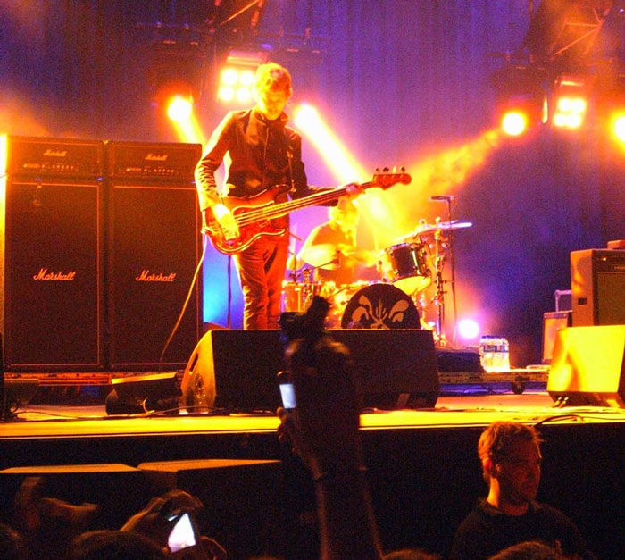 Musician playing bass guitar on stage with bright lights and Marshall amplifiers during a live concert performance.