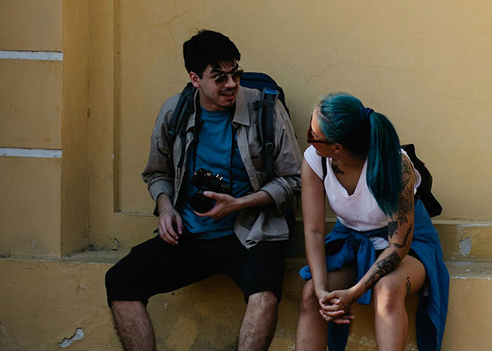 Two young travelers with backpacks casually talking while sitting against a yellow wall, capturing moments of coincidence.