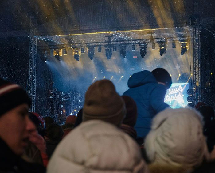 Crowd in warm clothing at an outdoor concert with stage lighting and fog highlighting the live music atmosphere.