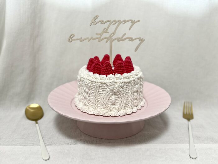 Crocheted realistic food-inspired birthday cake accessory with red strawberries on a pink plate by Japanese fiber artist.