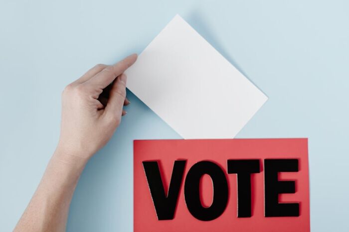 Hand placing a blank white card into a red box with bold black letters spelling vote, representing adulting skills.