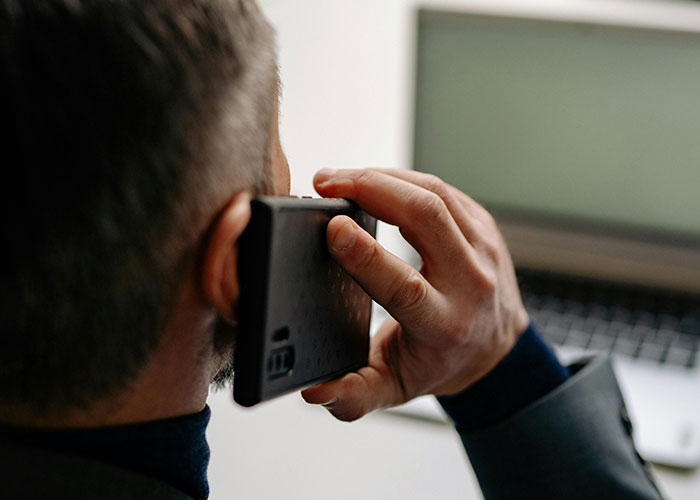 Man holding a phone to his ear while sitting near a laptop, illustrating a moment in criminal fails caught on camera.