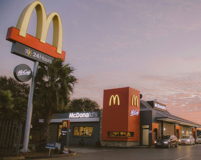 McDonald's restaurant at dusk with illuminated signs, a palm tree, and cars parked outside the entrance.