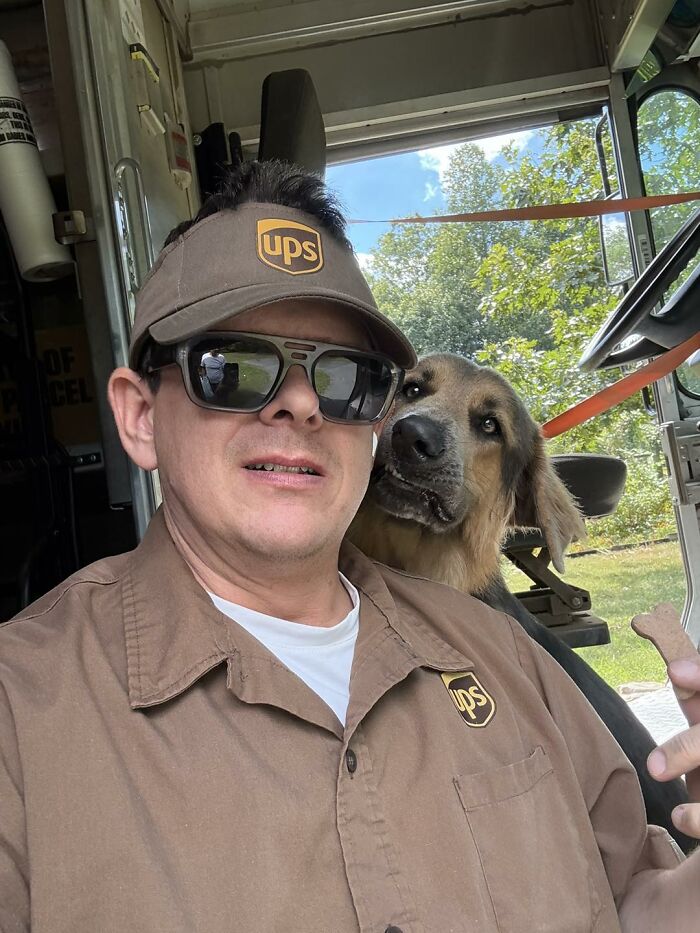UPS driver wearing uniform and sunglasses taking a selfie with an adorable dog inside a delivery truck on a sunny day.