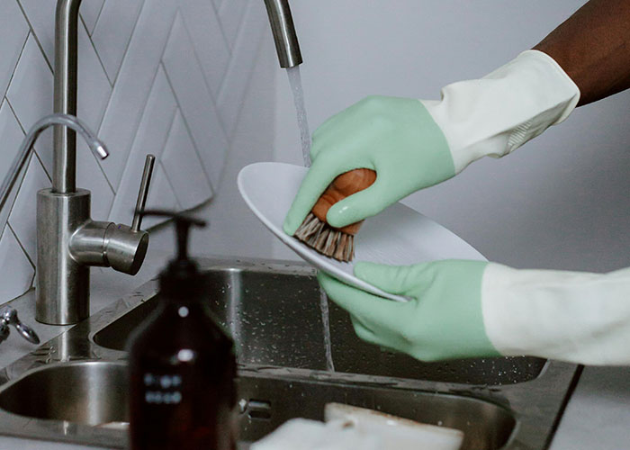 Person wearing gloves washing a dish in sink, illustrating common purchases people have ditched due to high price tags.