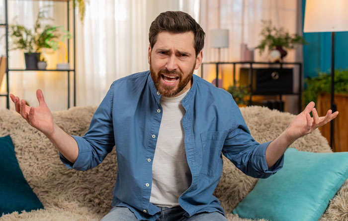 Frustrated man sitting on couch with arms open, expressing regret and tension in a cozy living room setting.