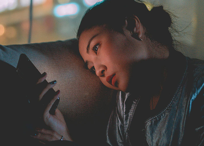 Young woman lying on a couch, looking thoughtfully at her smartphone, reflecting on historical events and societal damage.
