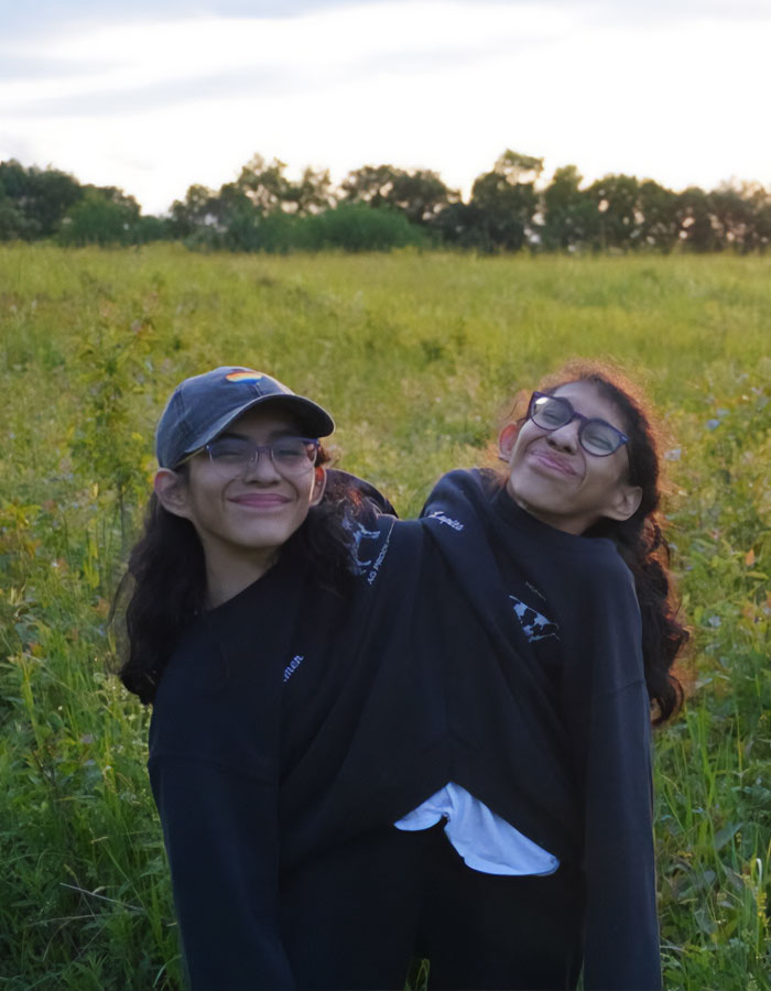 Conjoined twin Carmen Andrade smiling outdoors in a field, sharing a black sweatshirt, embracing natural surroundings.