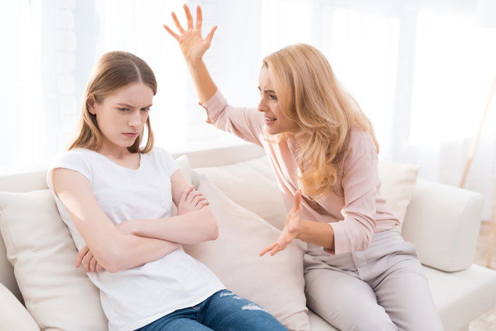 Woman telling wife why daughter doesn't trust, while daughter sits with arms crossed looking upset on the couch.