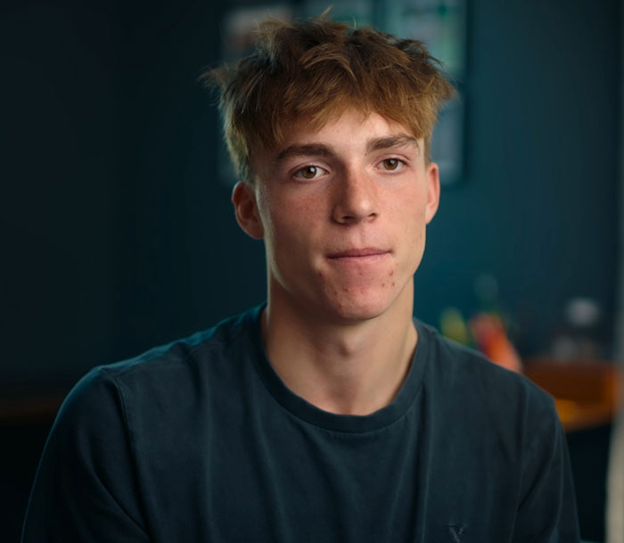 Teen boy with casual hair and dark shirt, looking thoughtful while sharing story about catfish torment and obsessed mom.