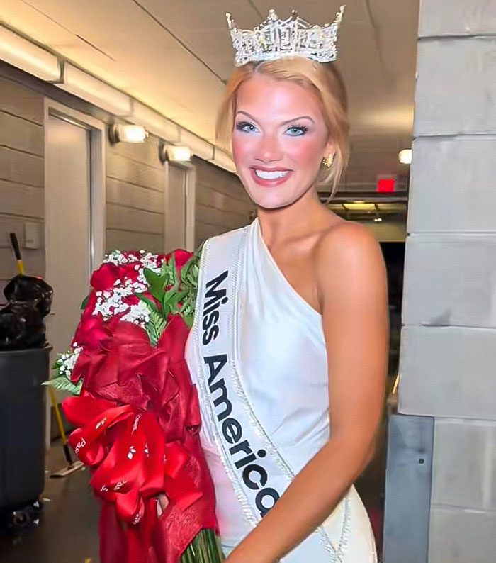 Newly crowned Miss America wearing crown and sash, holding bouquet, with notable makeup and concealer visible in hallway setting Newly crowned Miss America wearing crown and sash, holding bouquet, with notable makeup and concealer visible in hallway setting