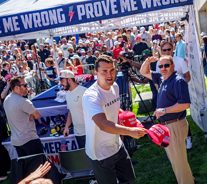 Charlie Kirk handing out Make America Great Again hats at a crowded outdoor political event with supporters around.