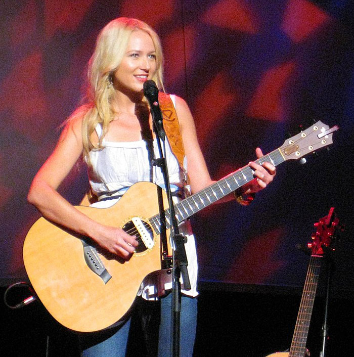 Female 90s star playing guitar on stage, smiling and engaging with fans, showcasing her toned body and fitness secrets.