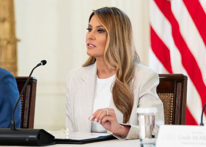 Melania Trump in a white blazer speaking at a formal event with an American flag in the background. Melania Trump in a white blazer speaking at a formal event with an American flag in the background.