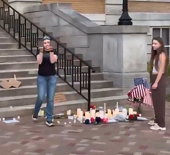 Two sisters at the site of a Charlie Kirk memorial with candles and flags, related to destruction and homelessness issues.