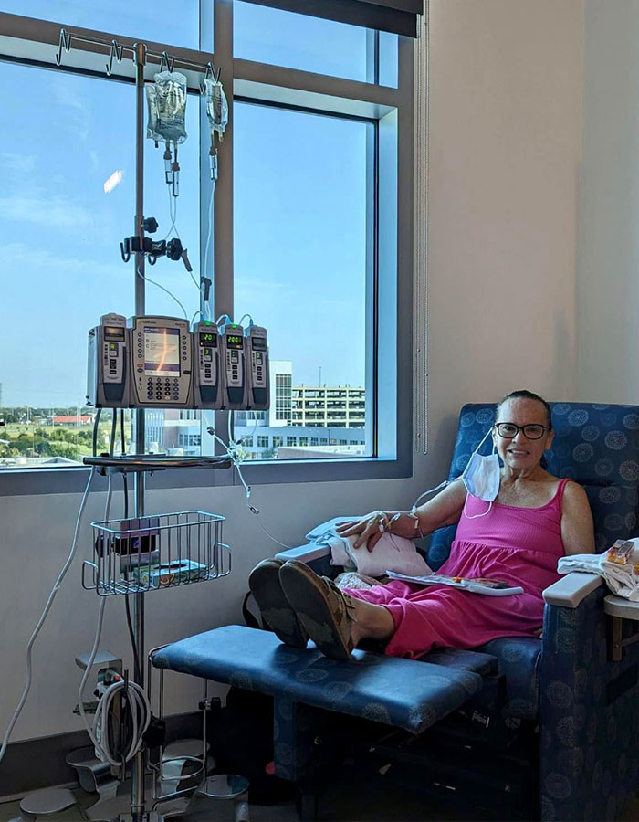 Woman receiving cancer treatment in hospital chair with IV drip beside large window showing cityscape outside.