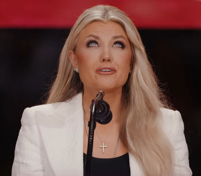 Woman in a white blazer wearing a cross necklace speaking into a microphone in context of Elon Musk hypocrisy news.