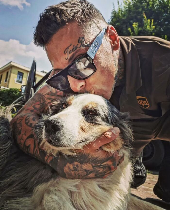 UPS driver with sunglasses and tattoos lovingly hugging and kissing an adorable dog during delivery with pets outdoors.