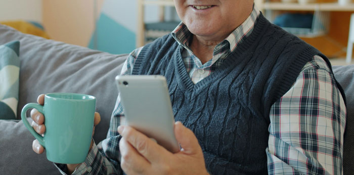 Hombre mayor sentado en sofá usando teléfono móvil mientras sostiene una taza, ilustrando cómo los teléfonos revelan verdades ocultas.