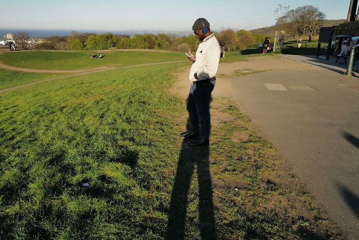 Man wearing headphones and sunglasses standing on grass with long shadow, captured in a brilliant street photo showing life’s funny side.