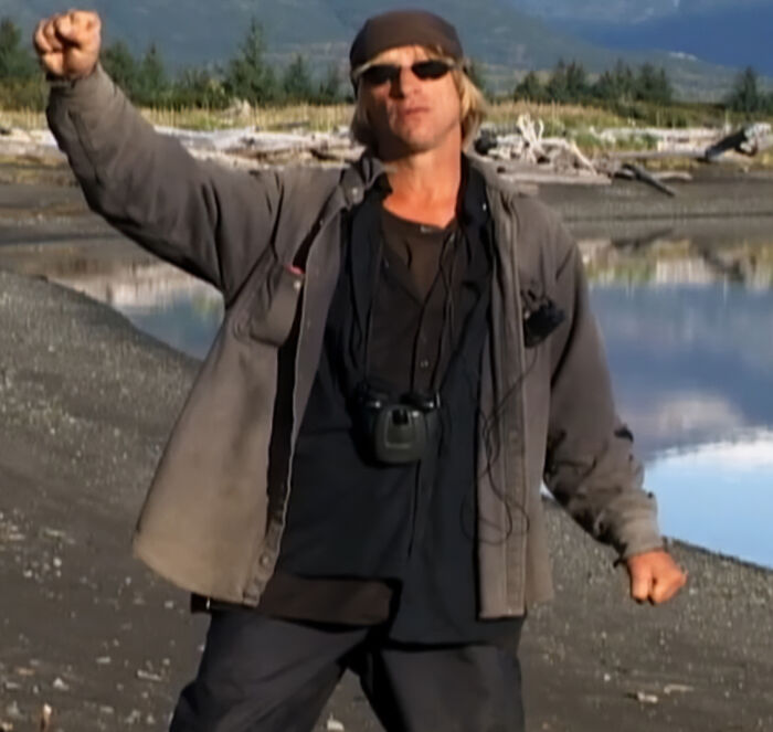 Man wearing sunglasses and outdoor clothing, raising fist near riverbank, related to famous people animal attacks topic.