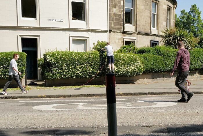 Child standing on a street bollard holding a dog leash while two pedestrians walk nearby in bright daylight street photos.