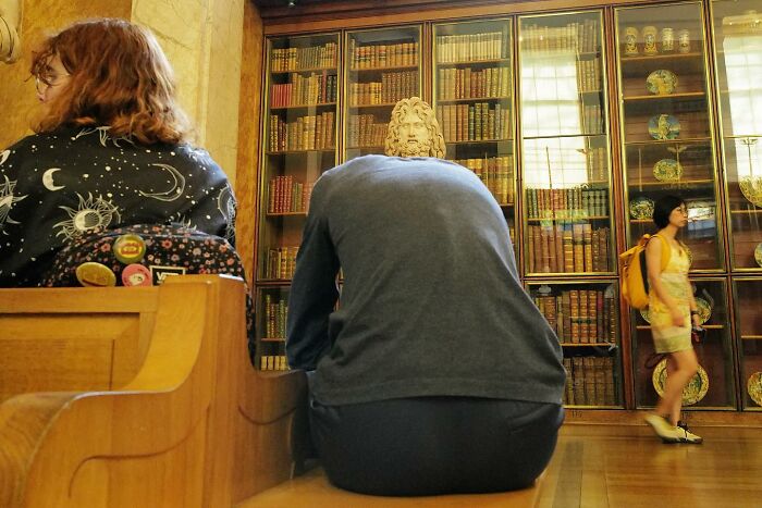 Person sitting on a bench with their back to a bust statue in a library, showcasing a brilliant street photo capturing the funny side.