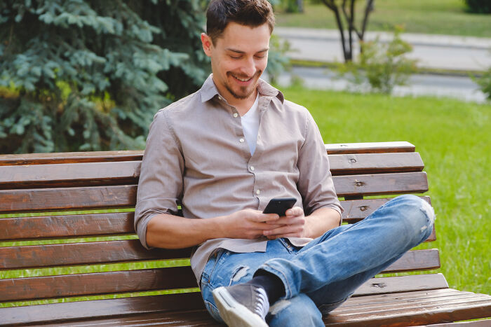 Young man sitting on a park bench smiling at his phone, illustrating absurd dealbreakers in relationships revealed by women.