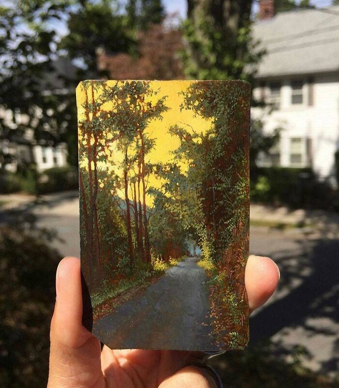 Hand holding a detailed painting by Dina Brodsky depicting a forest road with trees under a yellow sky.