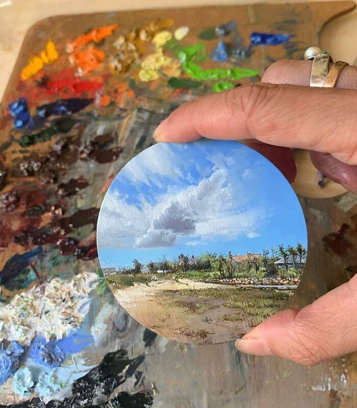 Hand holding a detailed miniature painting by Dina Brodsky depicting a natural landscape with blue sky and greenery.