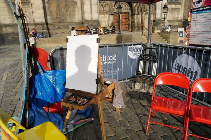 Street photo capturing a humorous shadow silhouette on a blank canvas in an outdoor urban setting with red chairs nearby.