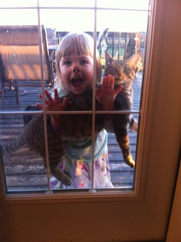 Young child holding a cat against a glass door, a wholesome post capturing a joyful and playful moment outdoors.