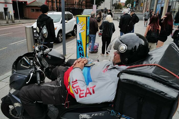 Delivery rider in helmet taking a break on a scooter parked on a busy urban street, street photos showing funny life moments