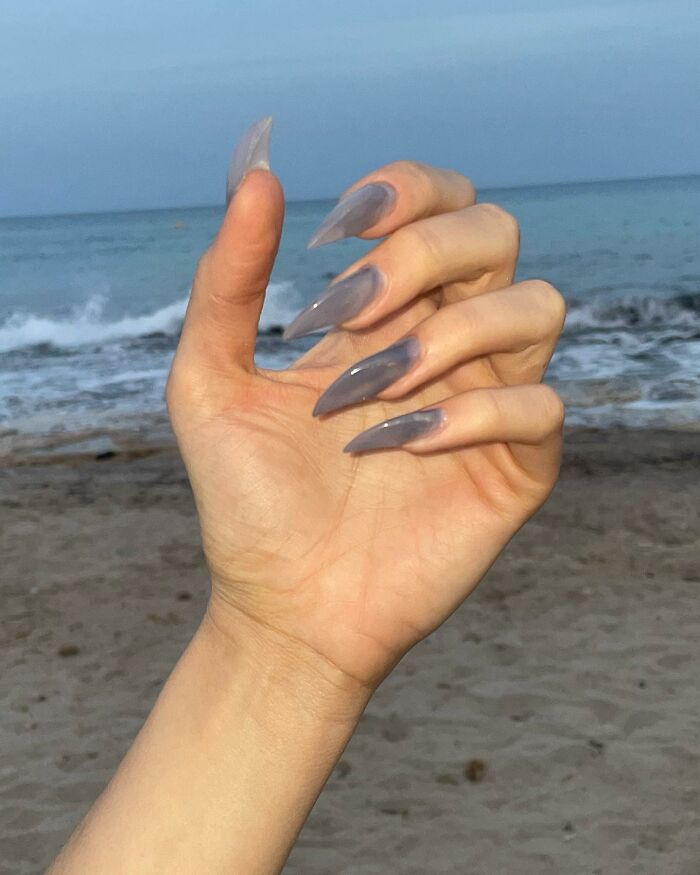 Hand with long, sharp stiletto nails in translucent grey nail designs posed in front of the beach and ocean background.