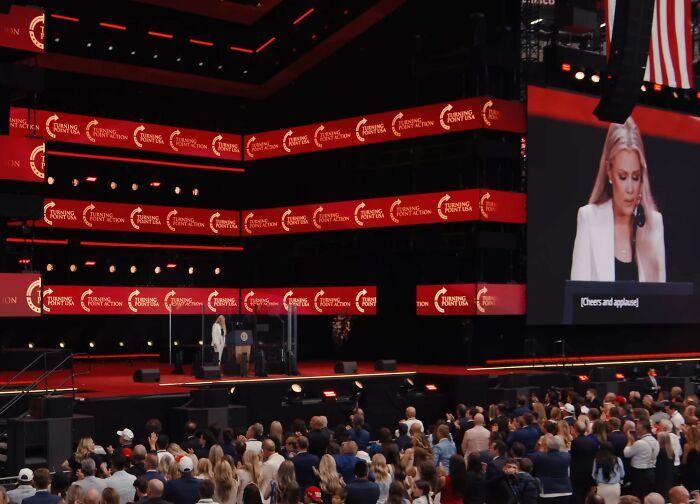 Woman speaking at a large event stage with crowd watching, amidst heated debate on Erika Kirk and feminist movement claims.