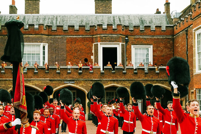 Guards in red uniforms raising hats in London courtyard, capturing atmosphere after the Queen’s passing.