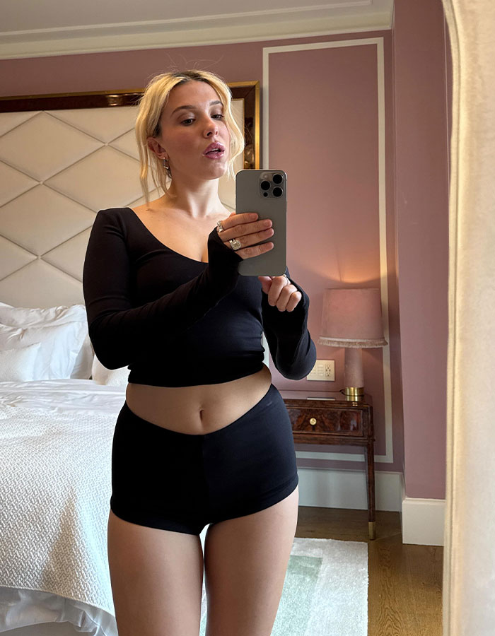 Woman taking a mirror selfie wearing a black pajama set, reflecting Millie Bobby Brown's post-mom style and appearance at home.