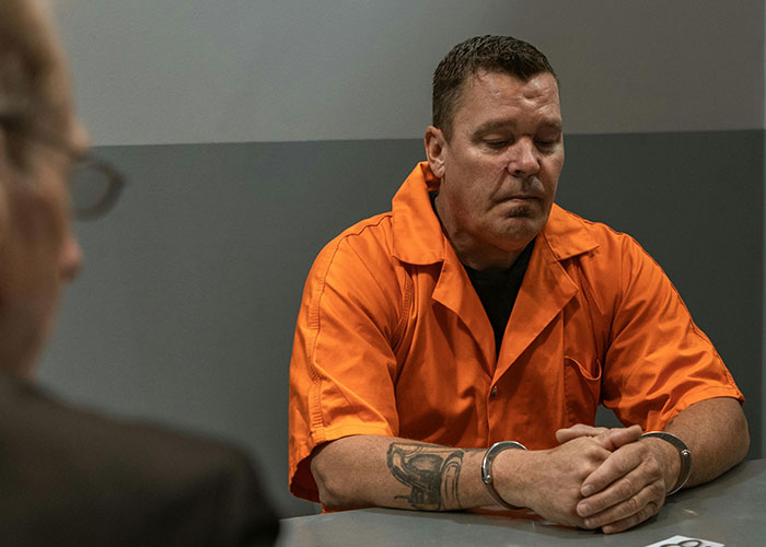 Man in an orange jumpsuit with handcuffs, sitting in an interrogation room, representing criminal fails humor.