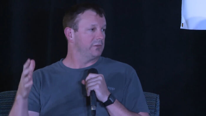 Man in a gray shirt holding a microphone, speaking about bold decisions with massive impact on the world.