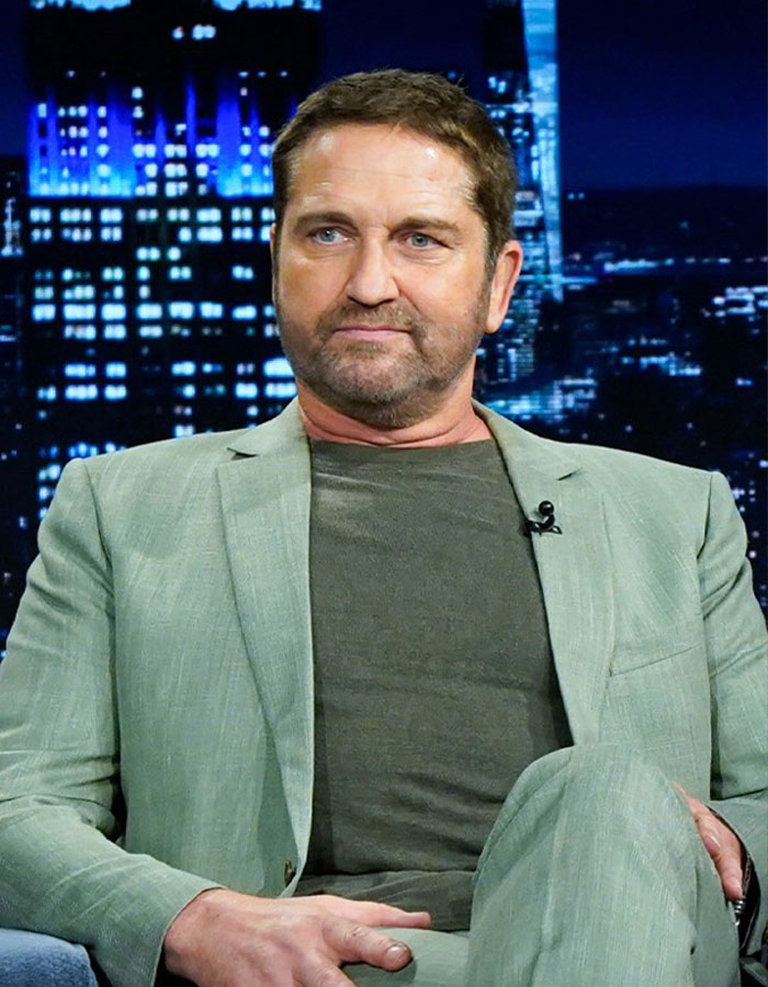 Man in a light green suit and dark shirt sitting in a studio with cityscape lights in the background, discussing Gal Gadot news.