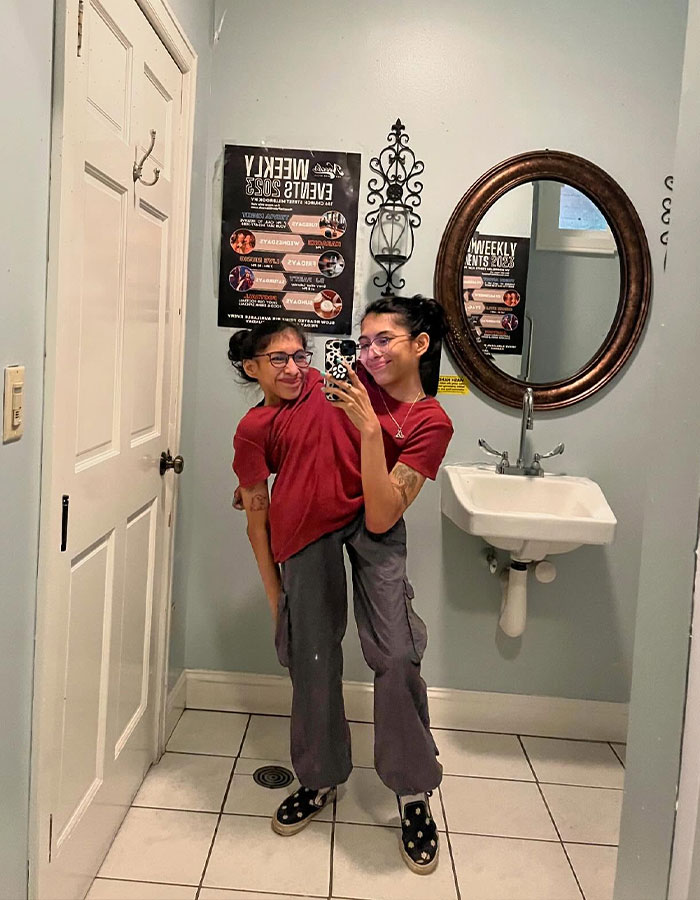Conjoined twin Carmen Andrade takes a mirror selfie smiling in casual clothes inside a tiled bathroom with a round mirror.