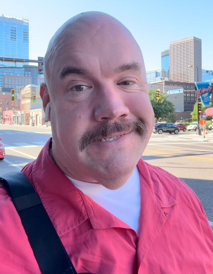 Man in red shirt outdoors with city buildings in background, relevant to passenger attacks plus-size seatmate too fat to fly topic. Man in red shirt outdoors with city buildings in background, relevant to passenger attacks plus-size seatmate too fat to fly topic.
