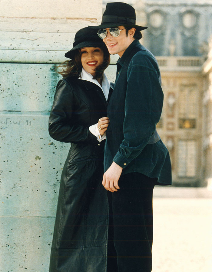 Michael Jackson and Lisa Marie Presley smiling outdoors, both wearing hats and dark-colored outfits in a historic setting.